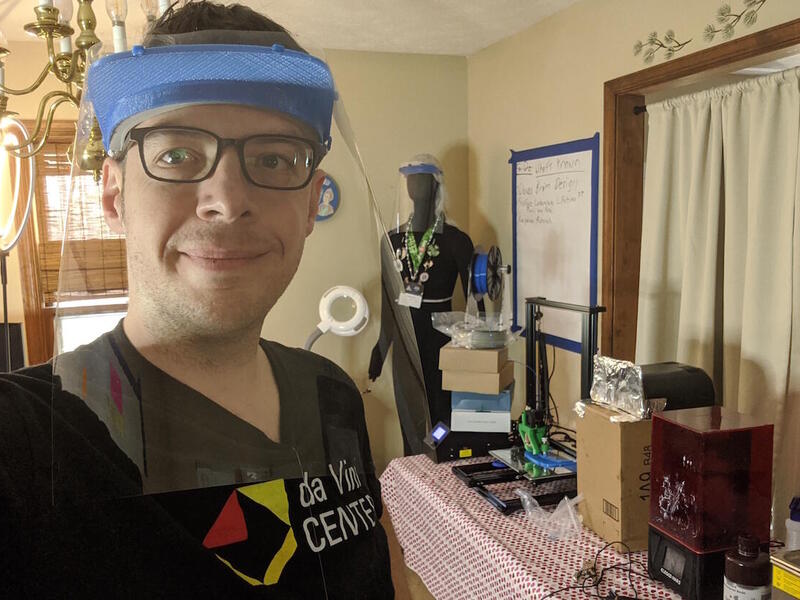 A student wearing a face shield stands in a room with equipment and various items.