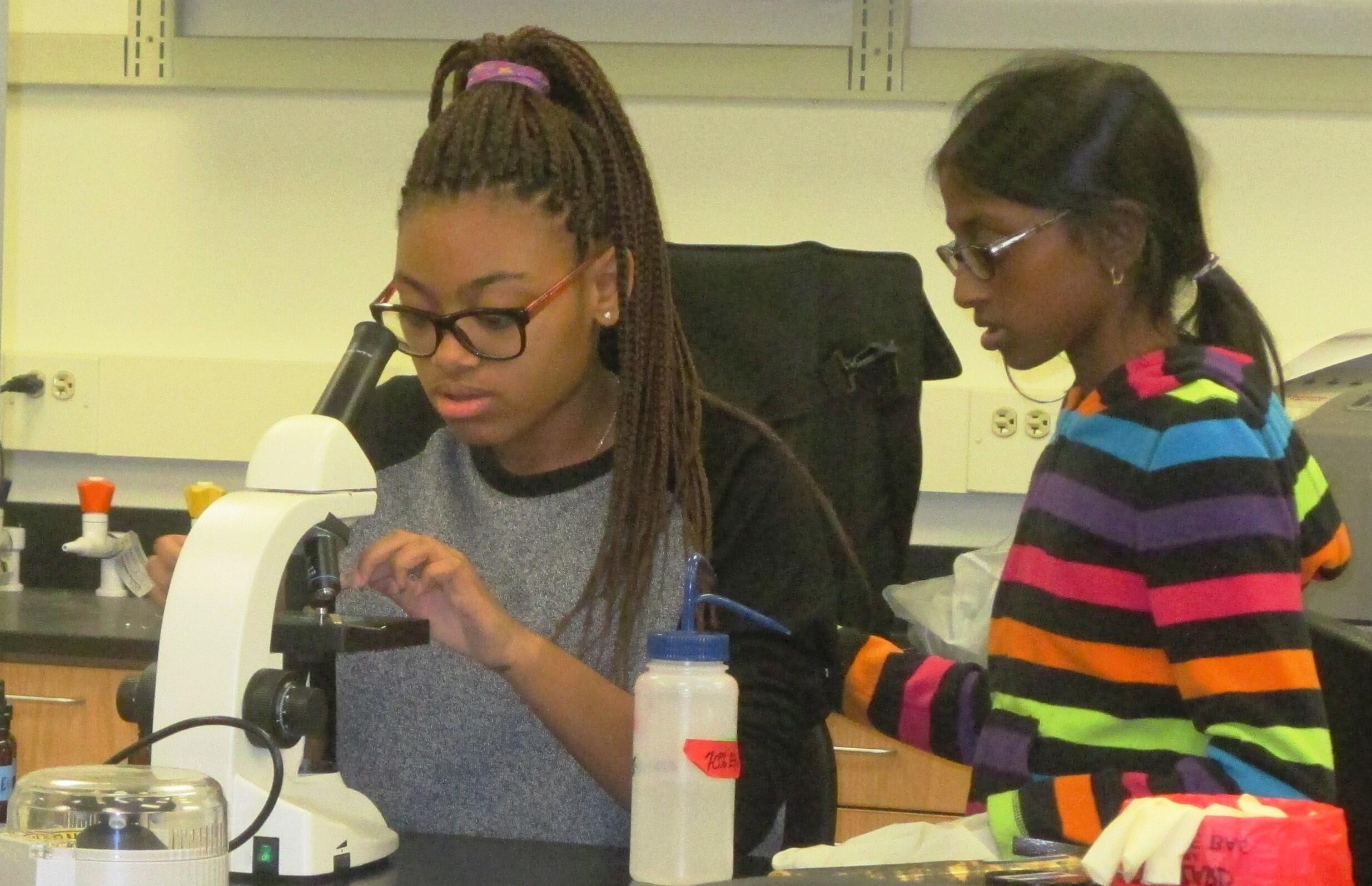 A woman looking into a microscope with a child standing next to her. 