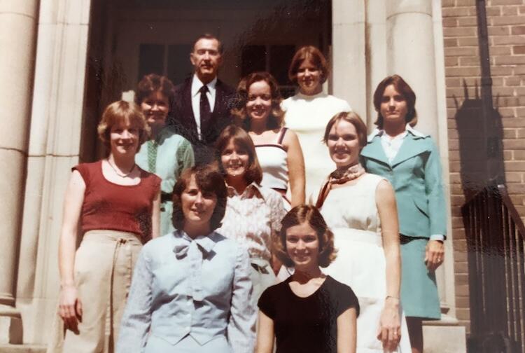 A group photo of nine women and one man standing on the stairs in front of a building. 