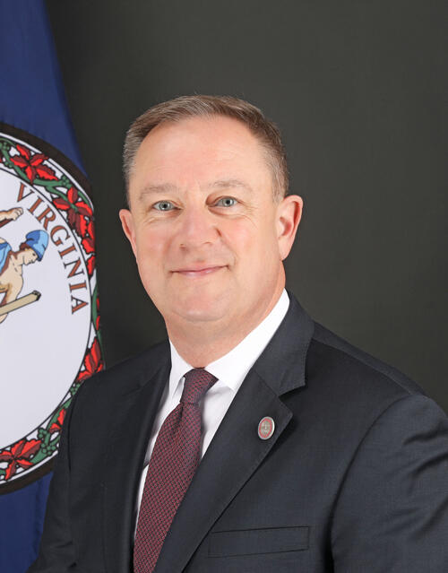 A photo of a man from the chest up. He is sitting next to a Virginia State Flag and wearing a suit and tie. 