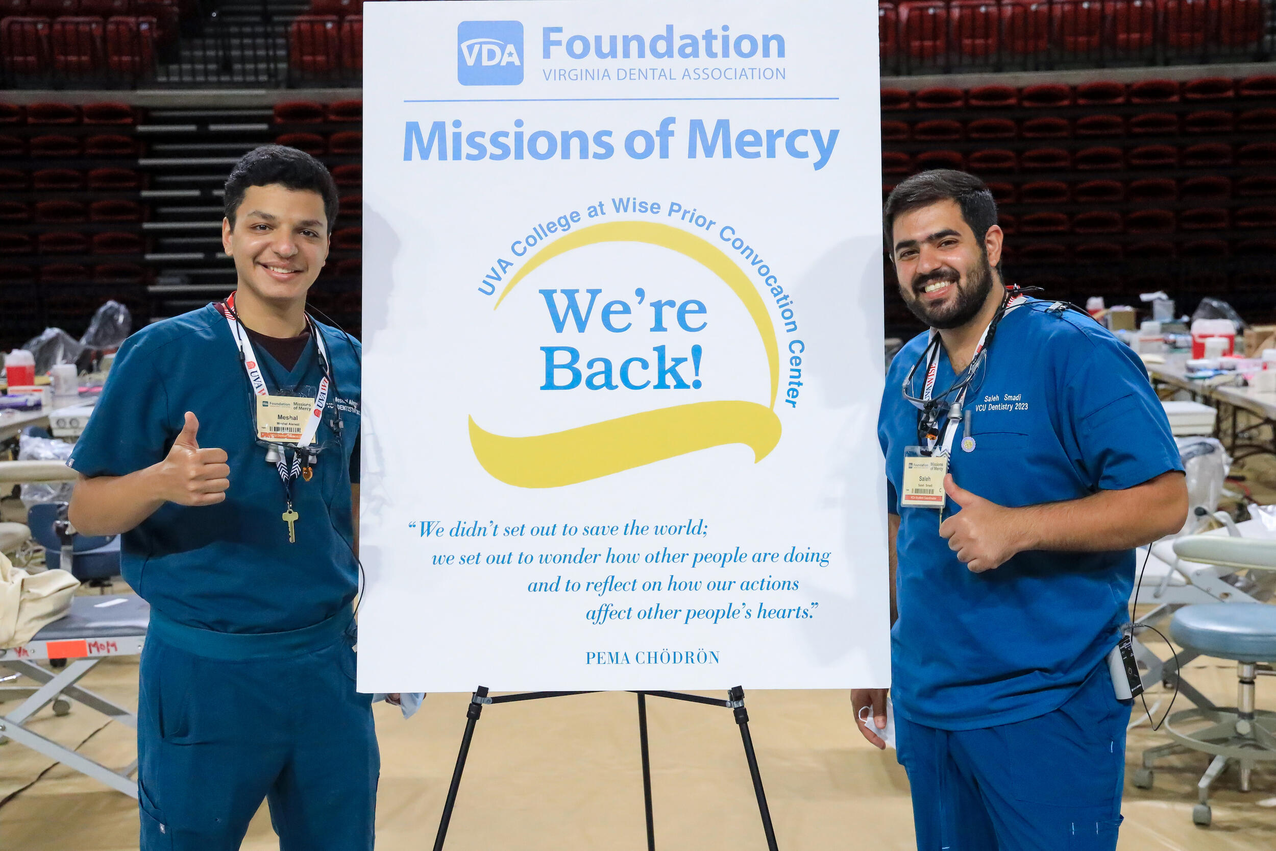 Two men on either side of a poster smiling with their thumbs up. The poster says \"Missions of Mercy We're Back\"