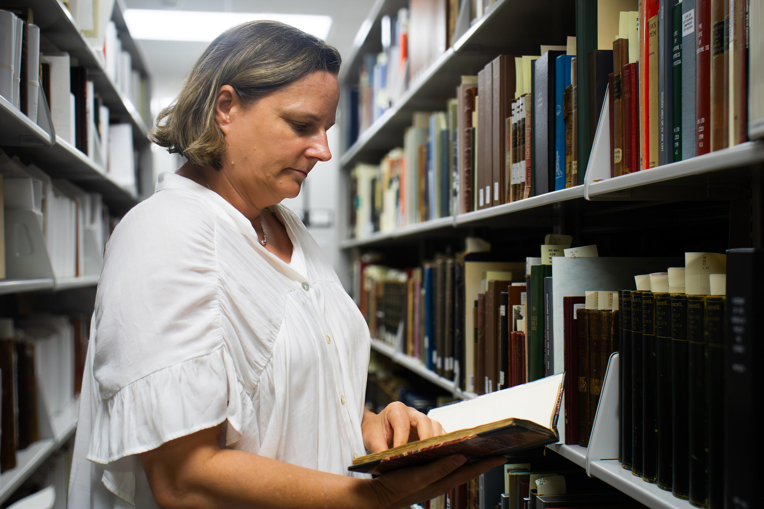 A photo of a woman from the waist up. She is reading a book in the middle of two aisles of books in a library.