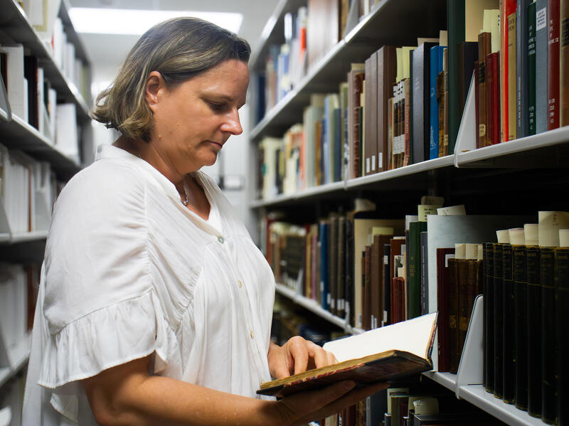 A photo of a woman from the waist up. She is reading a book in the middle of two aisles of books in a library. 