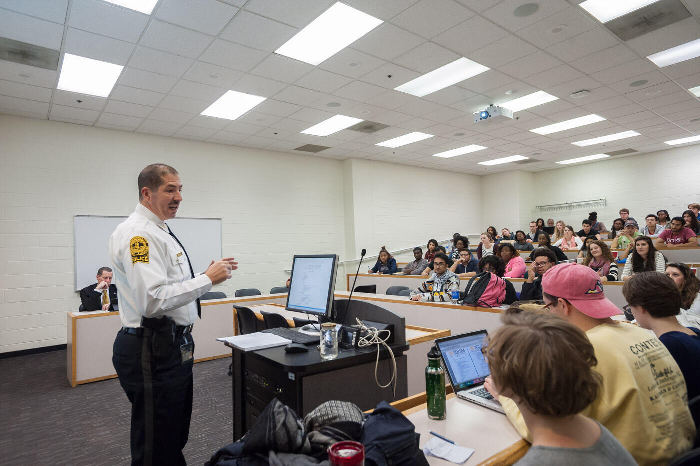 A photo of a man wearing a police uniform in a lecture hall standing behind a podium with a computer monitor and a microphone on it. In front of him are rows of seats.