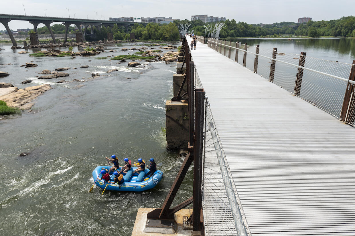 Pedestrians cross the T. Tyler Potterfield Memorial Bridge over the James River while a raft passes underneath.