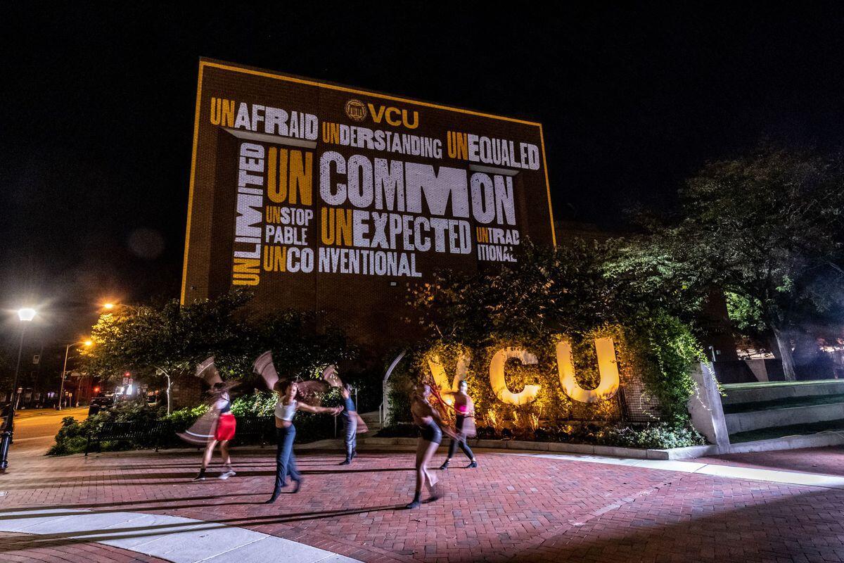 People walking across a plaza at night with lit up letters behind them that spell \"VCU.\" Above the letters is a large banner that says \"UNCOMMON\" in yellow and black text. 