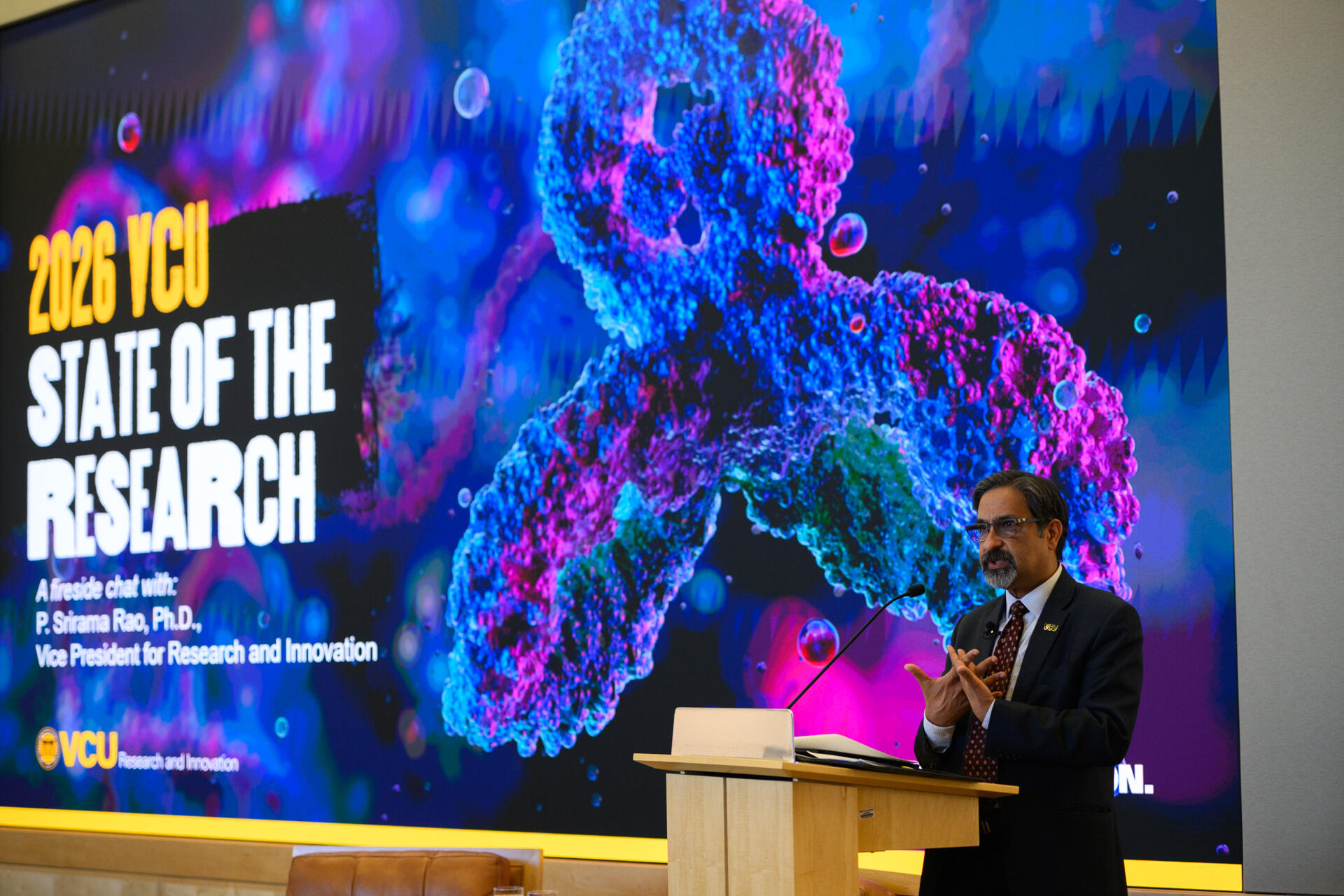 A photo of a man standing behind a podium and speaking. Behind the man is a screen that says \"2026 VCU STATE OF THE RESEARCH.\" 