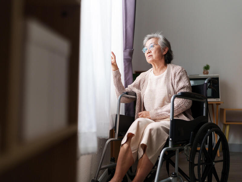 A photo of an older woman looking out a window while sitting in a wheel chair. 