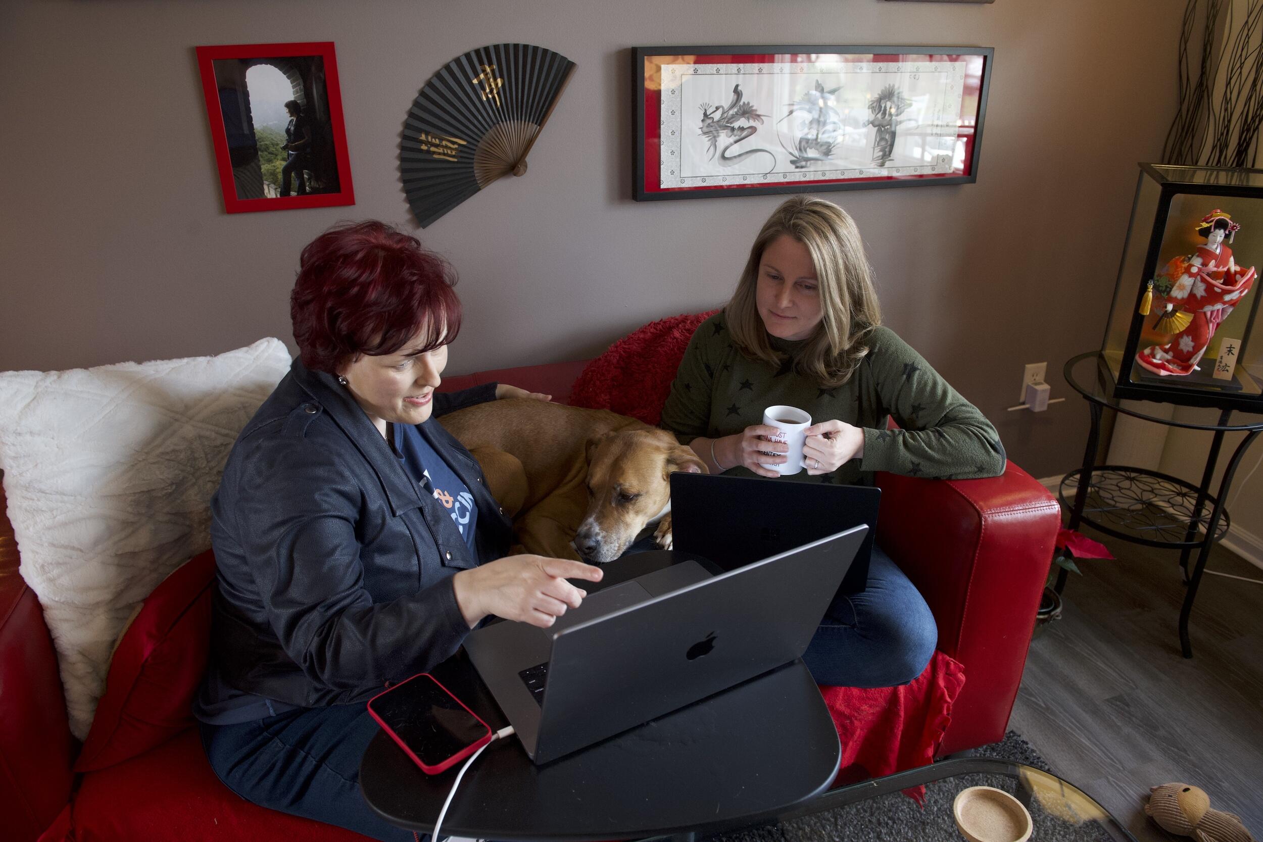 Two women sitting on a couch with a dog in between them. The woman on the right is holding a cup of coffee and the one of the left has a laptop in front of her that she is pointing at. Both women are looking at the laptop 