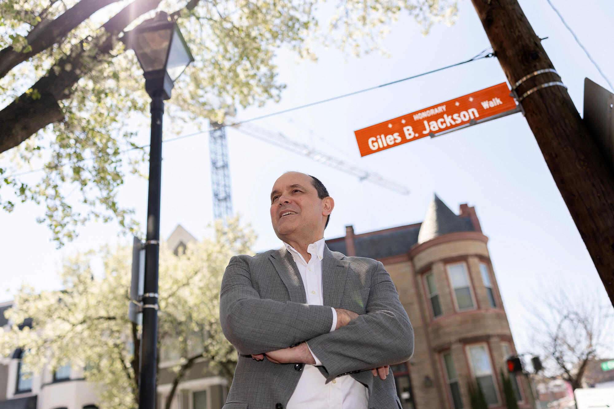 A photo of a man standing at a street corner with his arms crossed against his chest. 