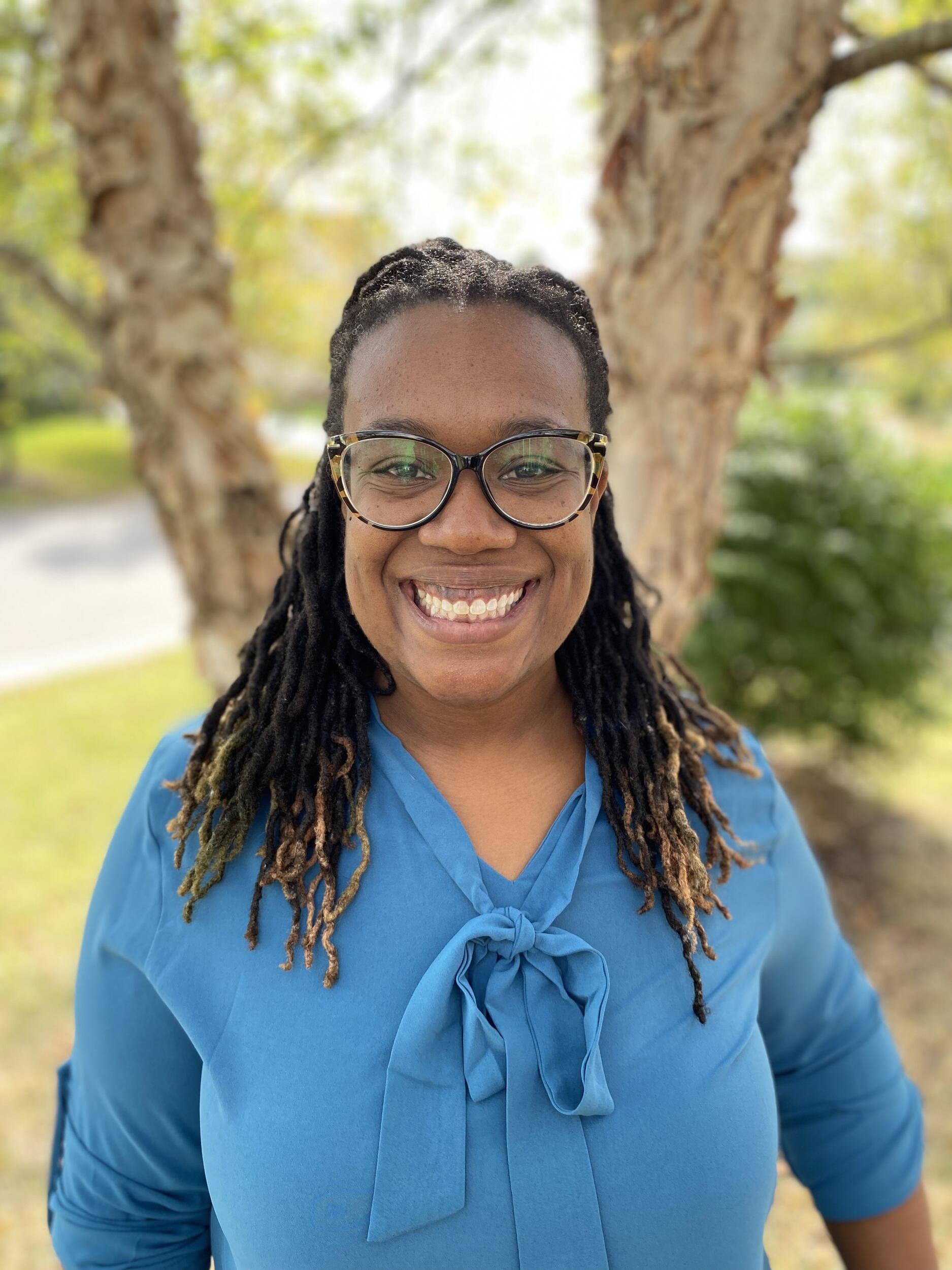 A photo of a woman wearing a teal blue shirt from the chest up. She is smiling and standing outside in front of a tree. 