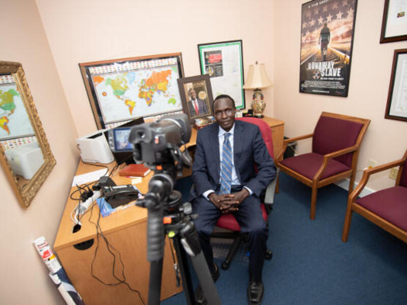 Man wearing a blue suit and blue striped tie seated by a desk in an office. A video camera on a tripod is in front of him.