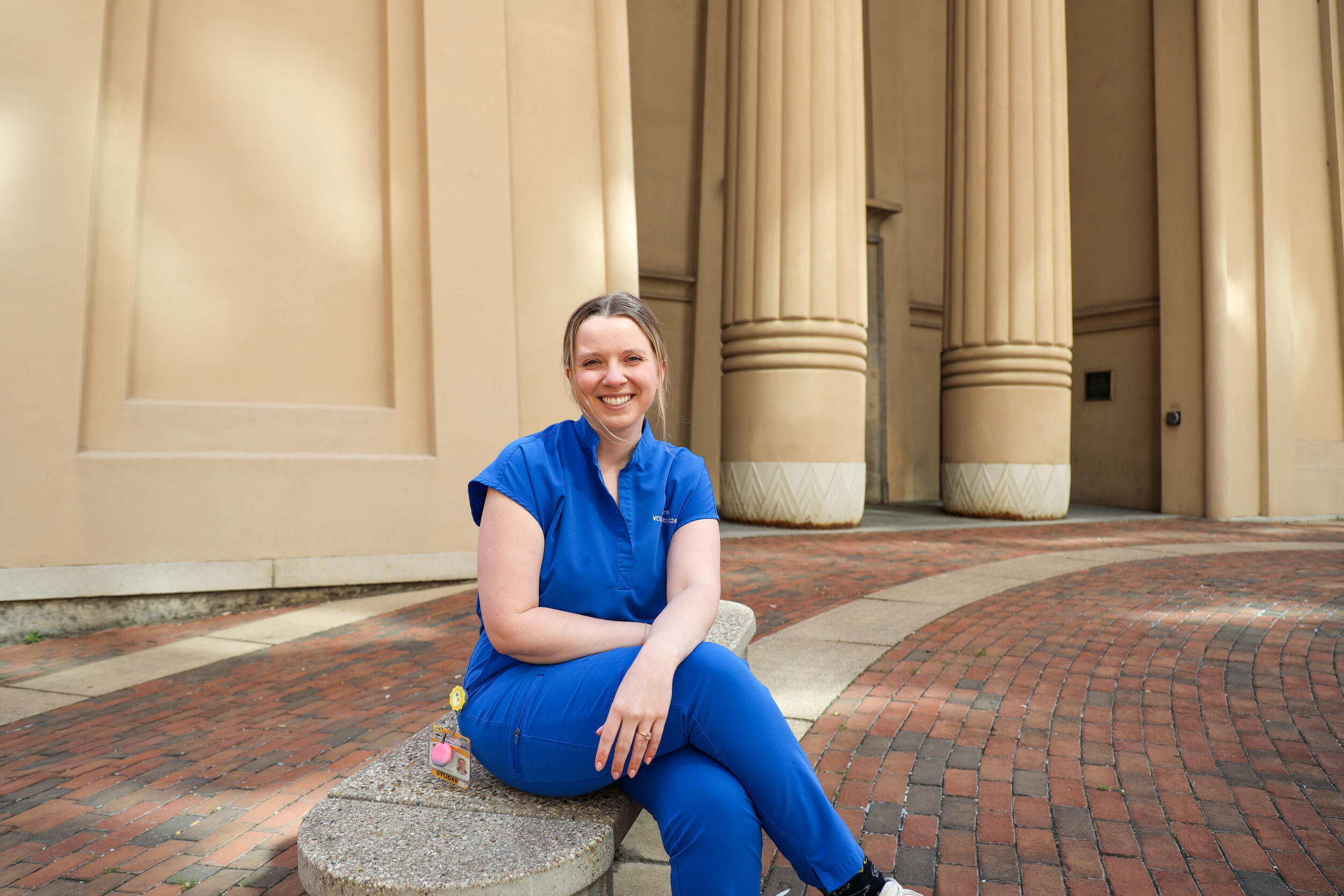 A woman in blue scrubs sits on a bench in front of a building with columns.