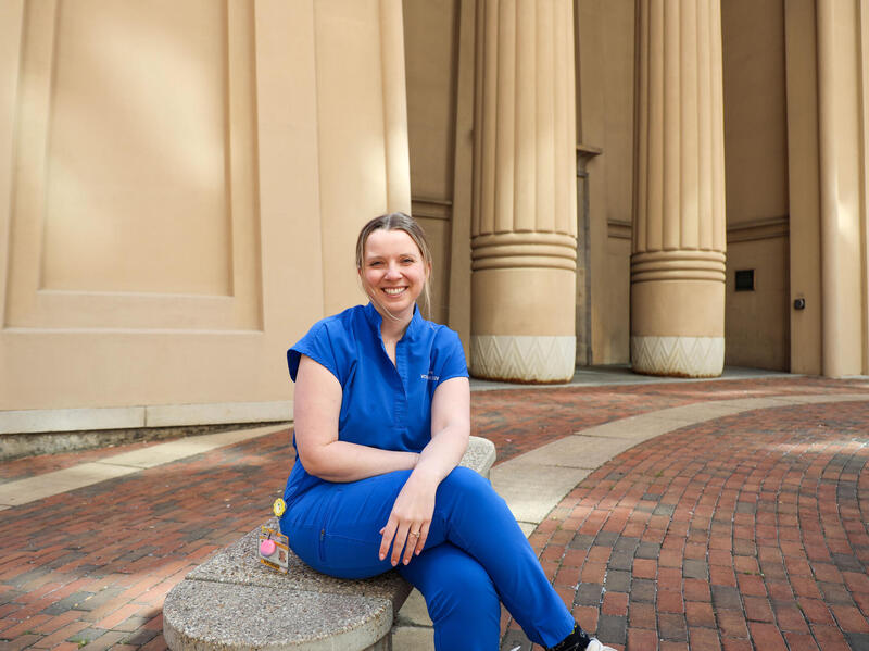 A woman in blue scrubs sits on a bench in front of a building with columns.