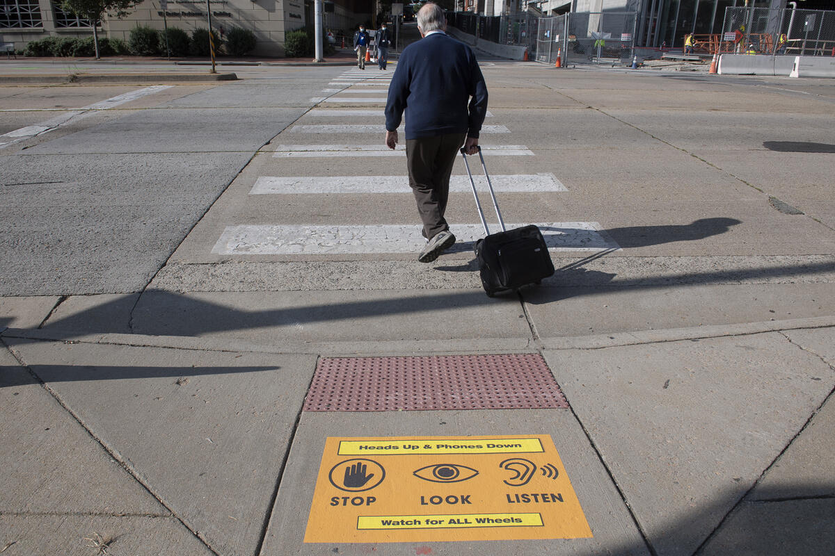 A pedestrian pulling a rolling briefcase begins to cross a street at a crosswalk. A sign placed on the sidewalk states \"Hands Up & Phones Down,\" symbols indicating pedestrians to \"STOP\" \"LOOK\" and \"LISTEN,\" and \"Watch for ALL Wheels.\"