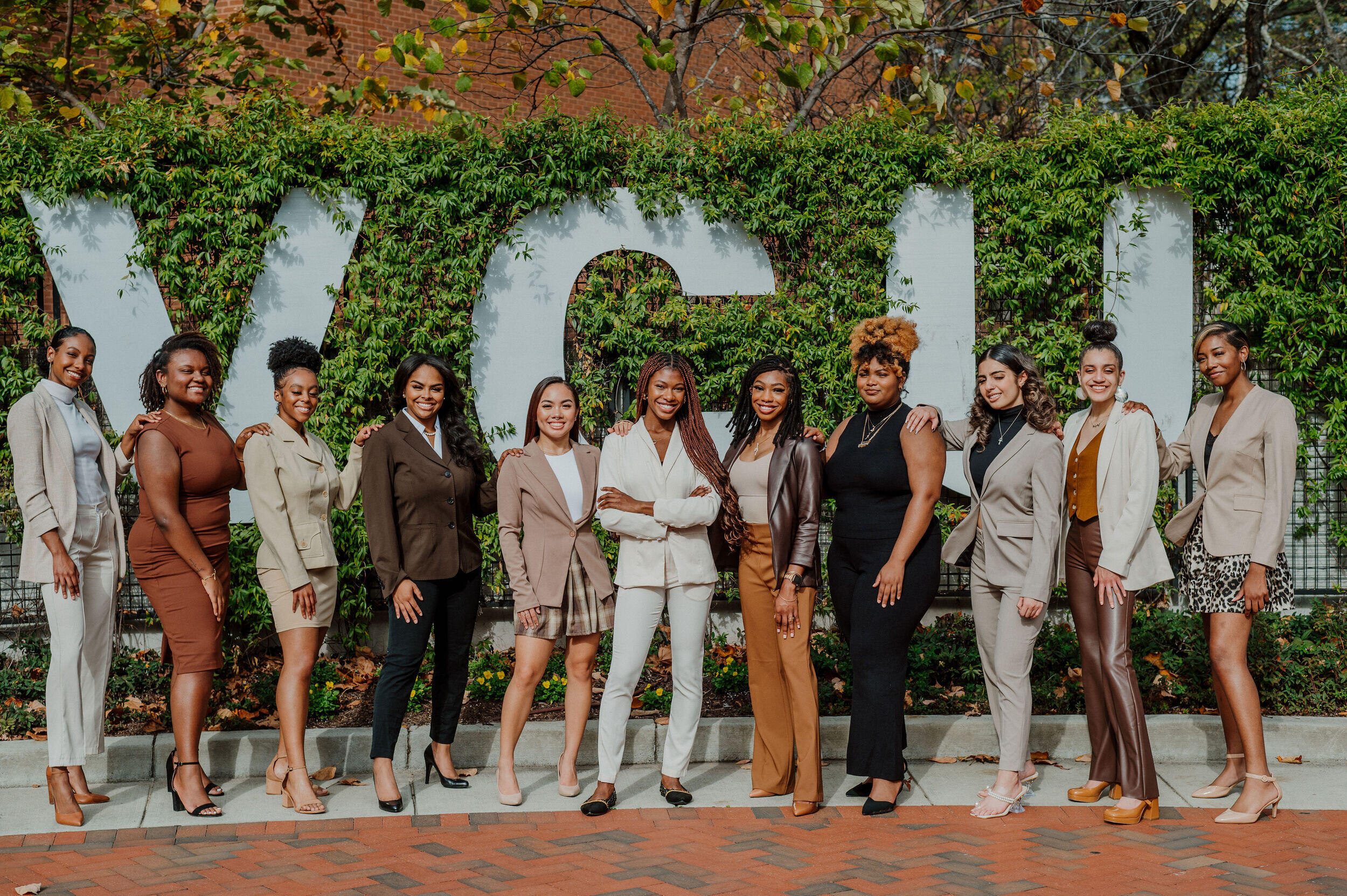 Eleven woman standing in front of a hedge with white letters that spell out \"VCU\"