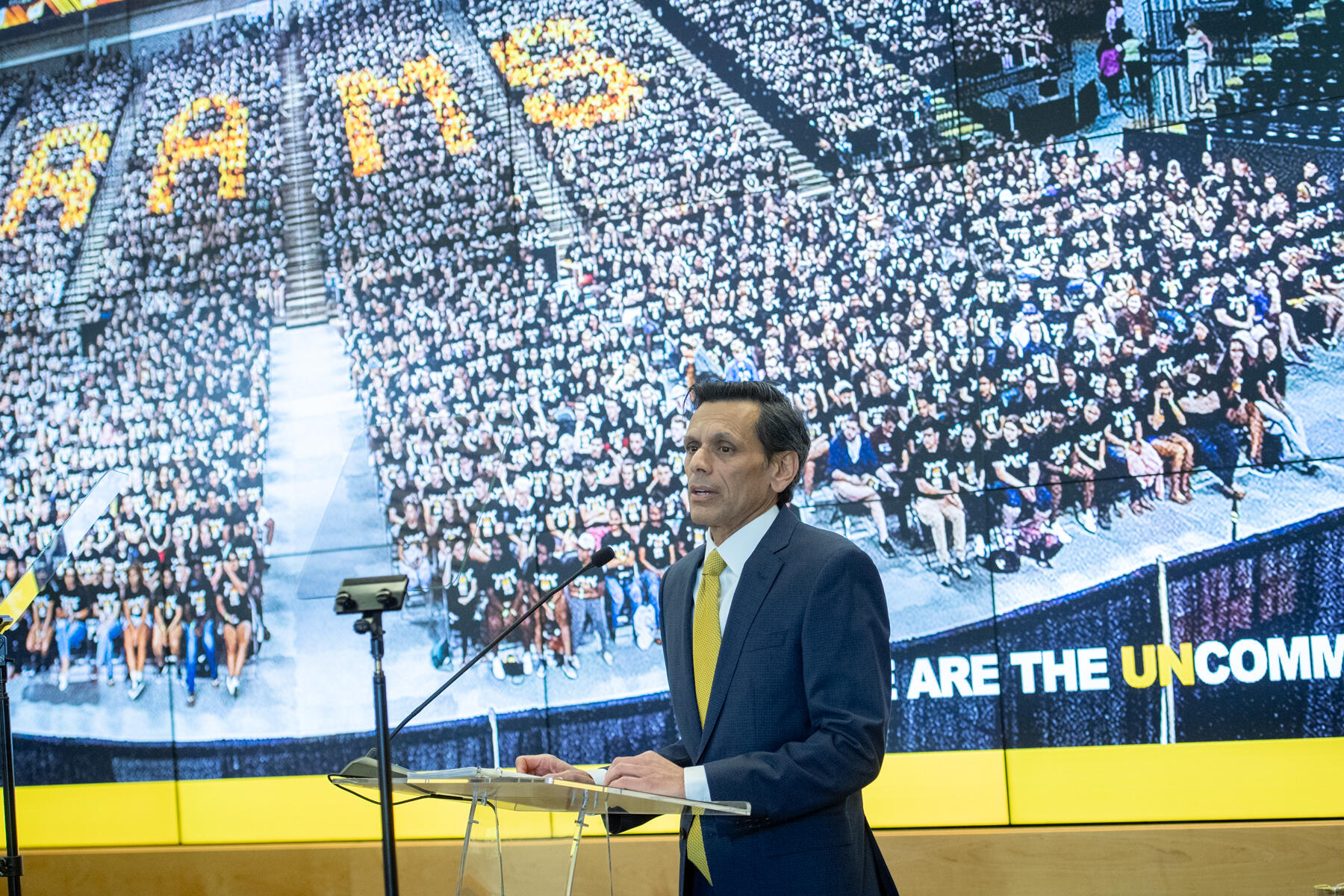 Photo of a man in a suit with a yellow tie speaking at a lectern in front of an image of a large crowd of sitting people.