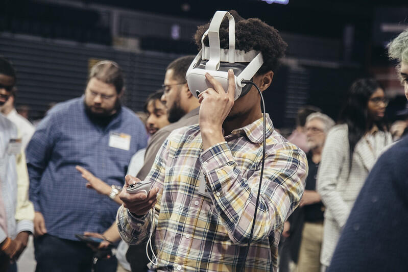A photo of a man wearing a VR headset and holding a remote. He is surrounded by other people who are walking and looking at other things. 
