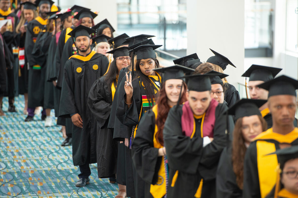 A photo of a line of students wearing graduation gaps and gowns in a line. One student is looking at the camera and has her hand up in a peace sign. 