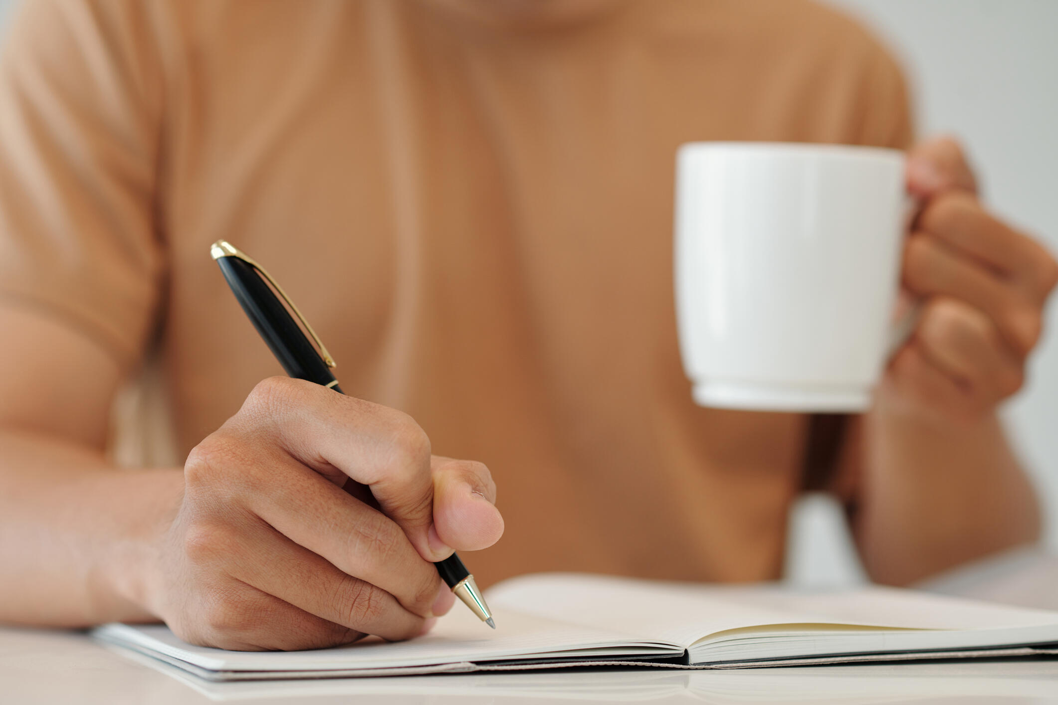 A photo of a man sitting at a table writing in a notebook while holding a cup of coffee.