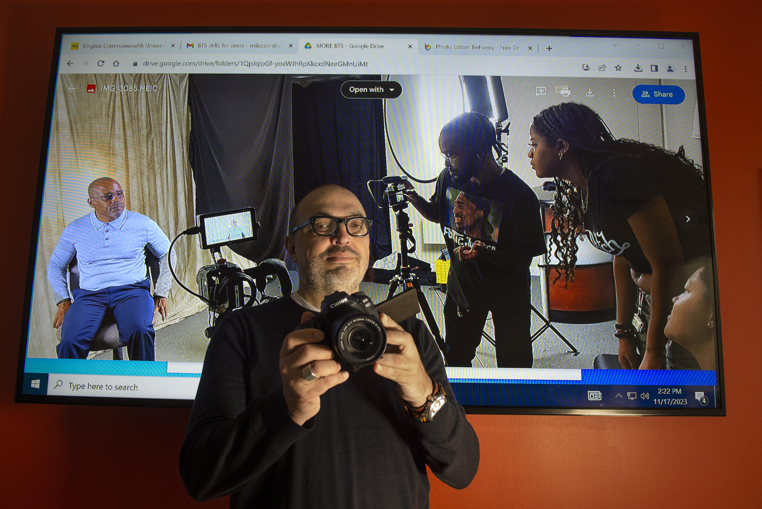 A photo of a man holding a video camera while standing in front of a large screen which shows two people filming another person on a set. 