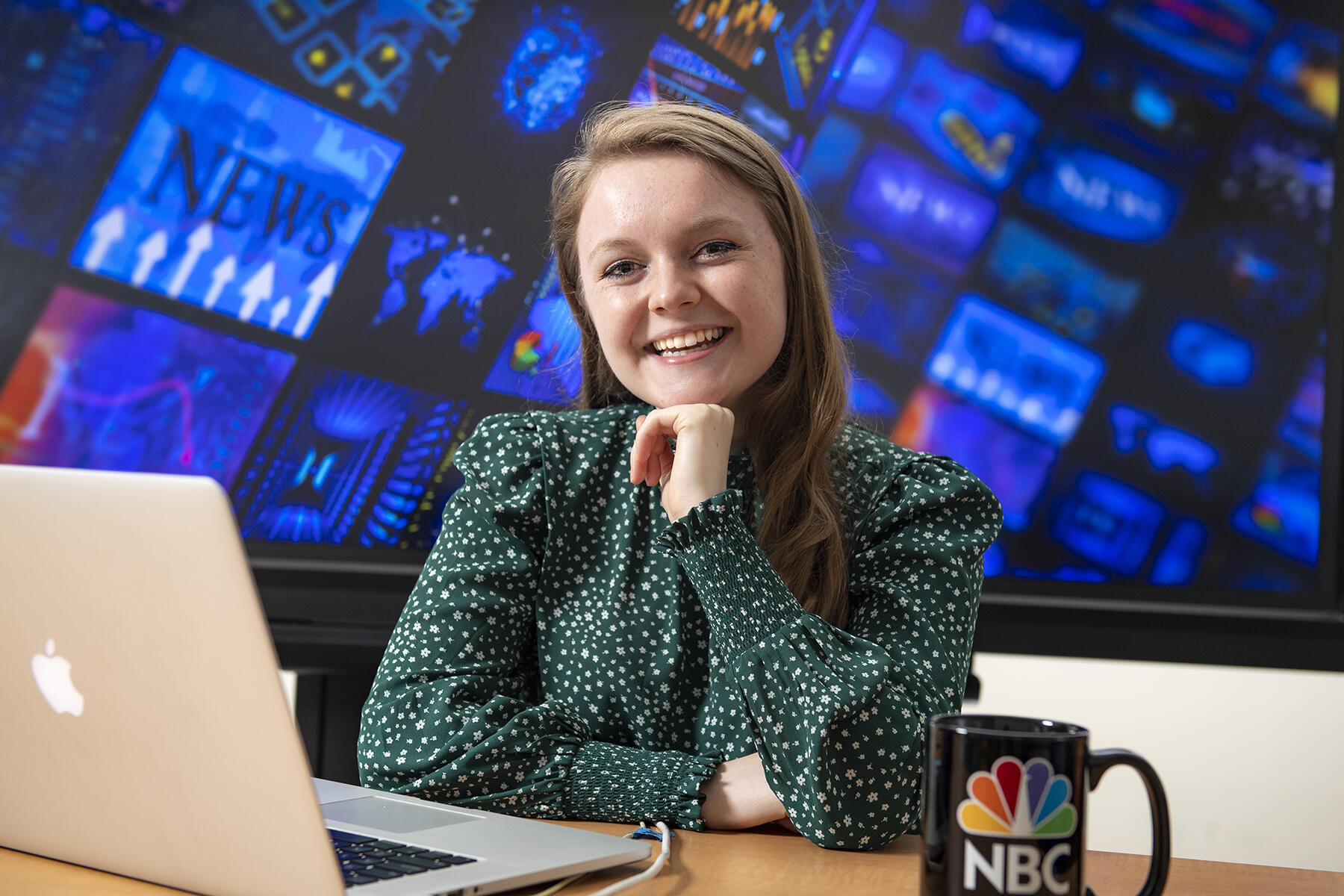 Allison Martin sitting at a desk with a laptop in front of her and a coffee mug that says \"NBC.\" 