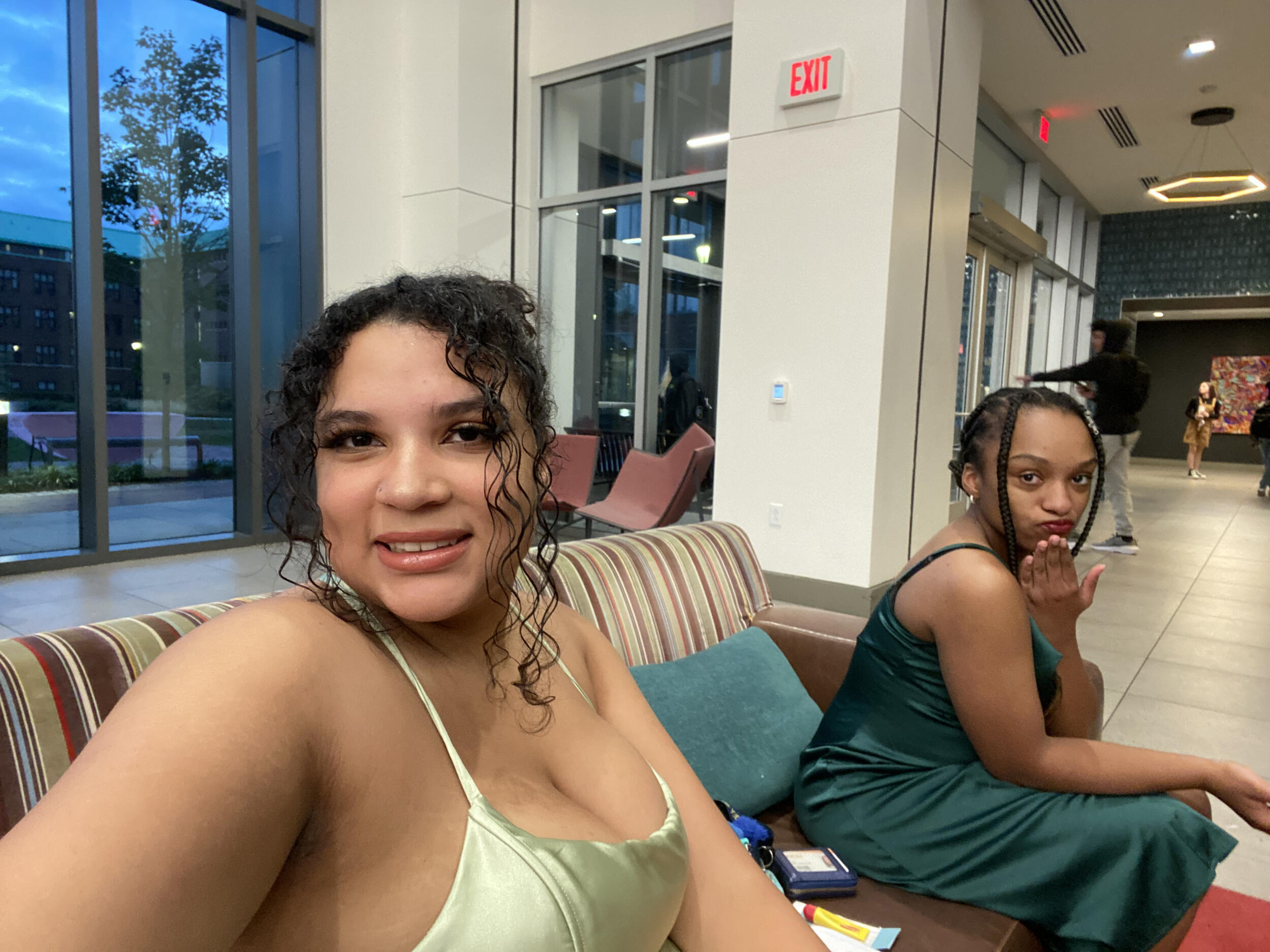 A photo of two women sitting on a couch. Both women are wearing formal dresses and have their hair tied up. 