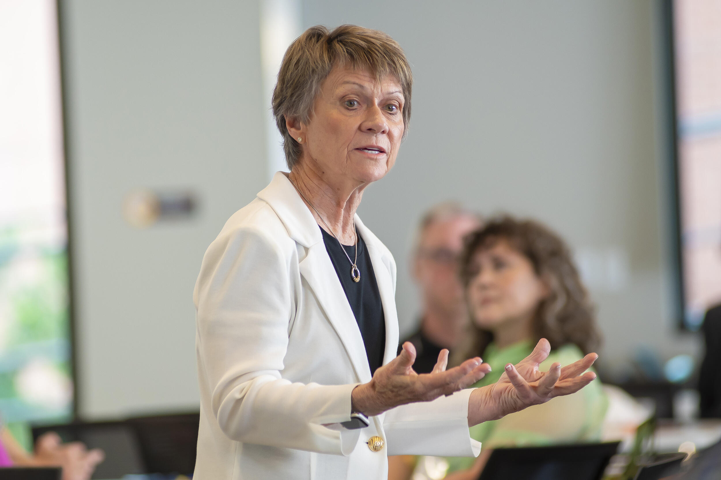A photo of a woman from the waist up. She is spekain in a lecture room and there are other people watching her speak. 