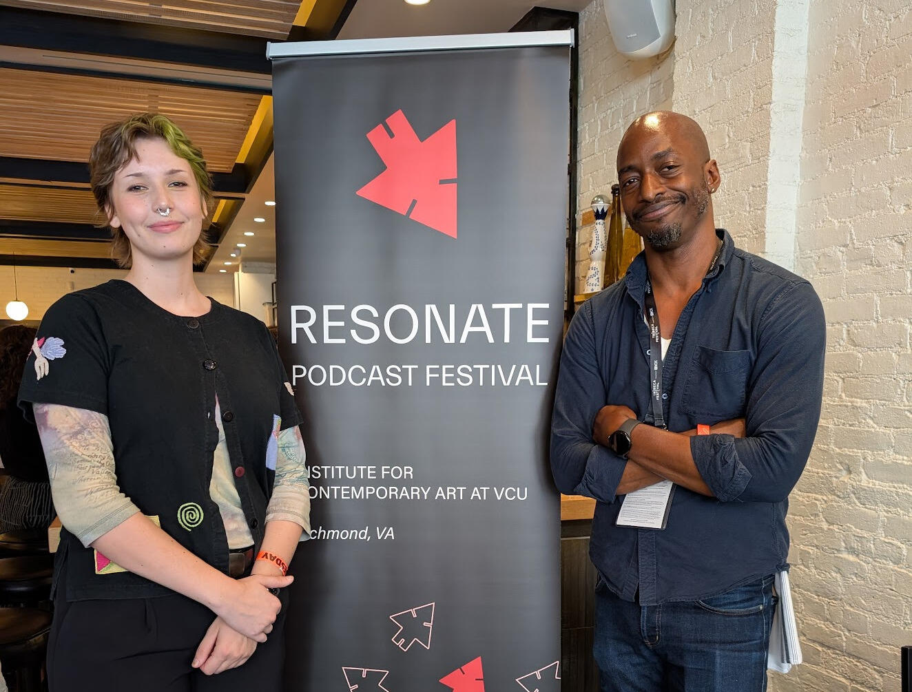 A photo of two people standing on either side of a black banner with white text that reads \"RESONATE\" \"PODCAST FESTIVA\" \"INSTITUT FOR CONTEM PORARY ART AT VCU\" \"Richmond, VA\"