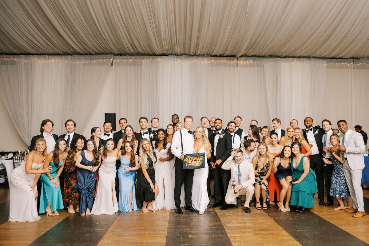 A group of people in formal wedding attire standing together. In the middle a bride and groom hold a black sign that says \"V C U\" in yellow letters:
