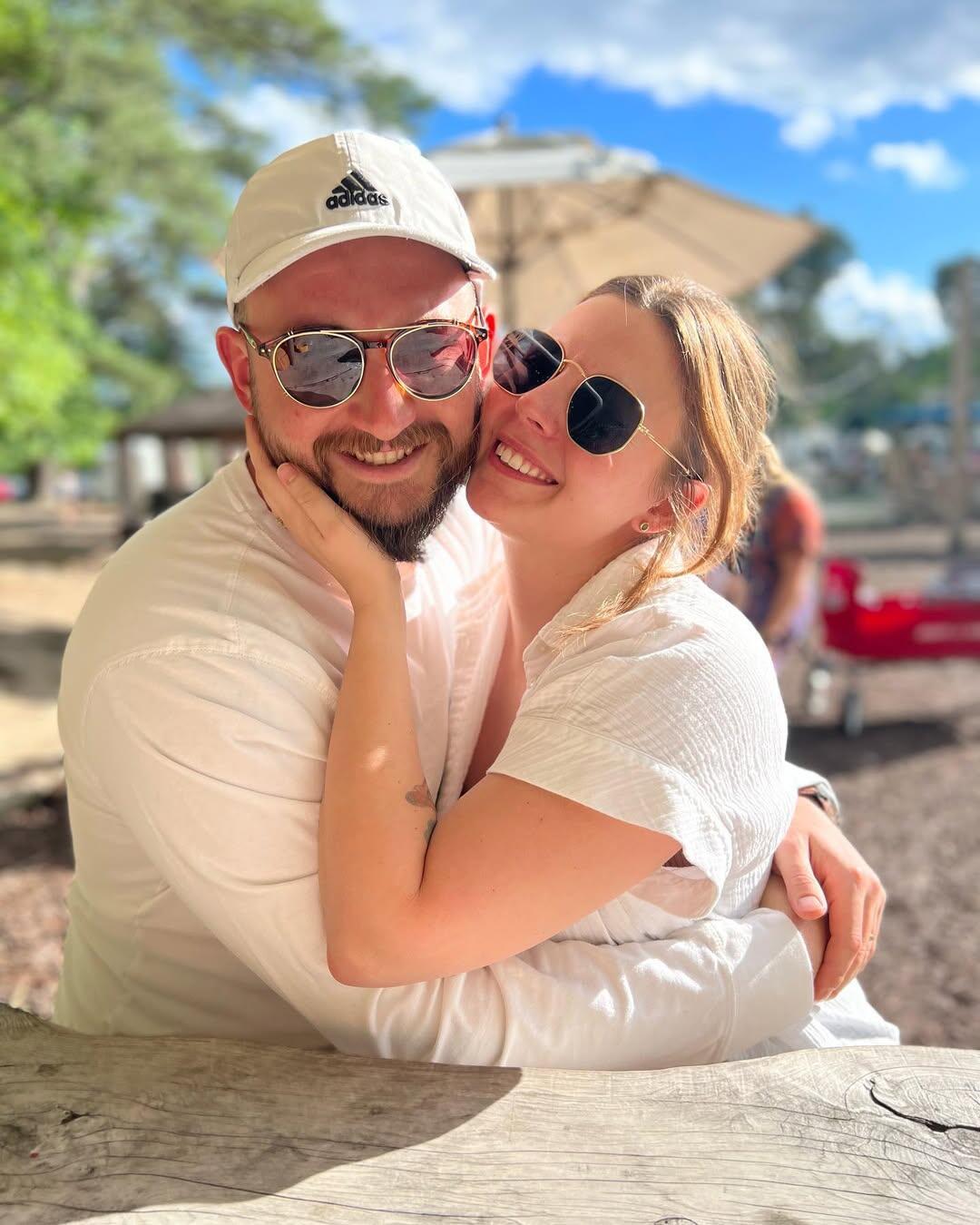 A photo of a man with his arms around a woman. The woman has her hand cupping the side of the man's face. Both of them are smiling and wearing sunglasses. 