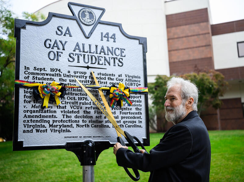 A photo of a man cutting yellow, black, and rainbow ribbons wrapped around a sign with a large pair of scissors. The top of the plaque says \"SA\" \"144\" \"GAY ALLIANCE OF STUDENTS.\"