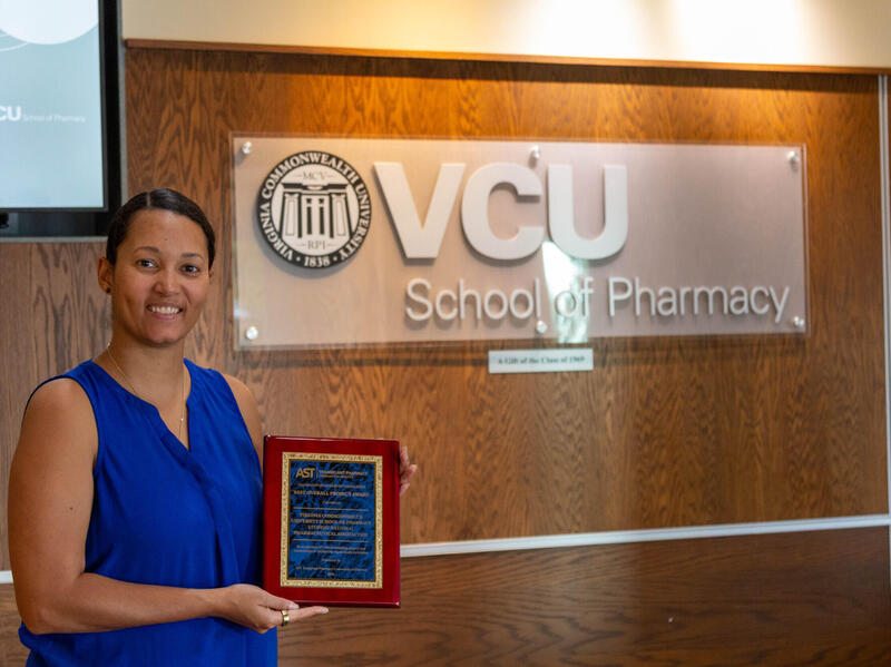A photo of a woman from the waist up. She is holding an award plaque. The woman is standing to the left of a sign that says \"VCU School of Pharmacy.\" 