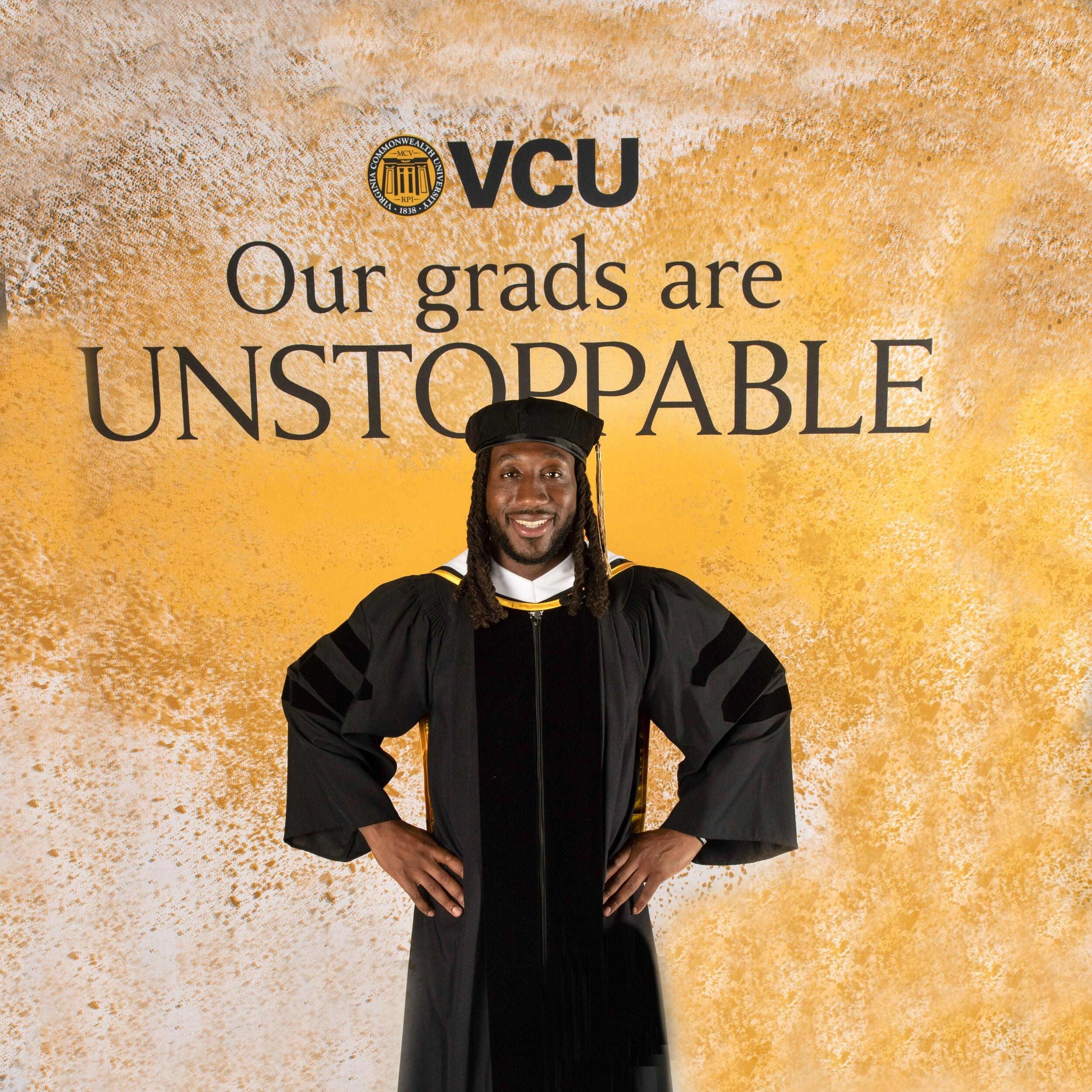 A photo of a man wearing a cap and gown standing with is hands on his hips. Behind him is black text that reads \"VCU Our grads are UNSTOPPABLE\"
