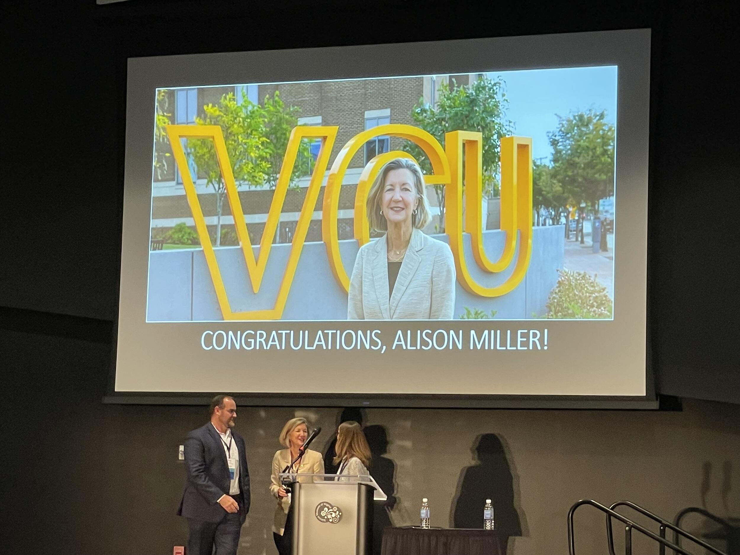 Three people standing on a stage in front of a projection screen. On the screen is a photo of a woman and white text that reads \"CONGRADULATIONS, ALISON MILLER!\" 