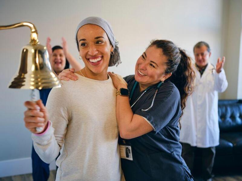 A photo of a woman smiling and ringing a bell. Next to her a nurse is holding onto her sholder. Behind them another man and woman are clapping. 