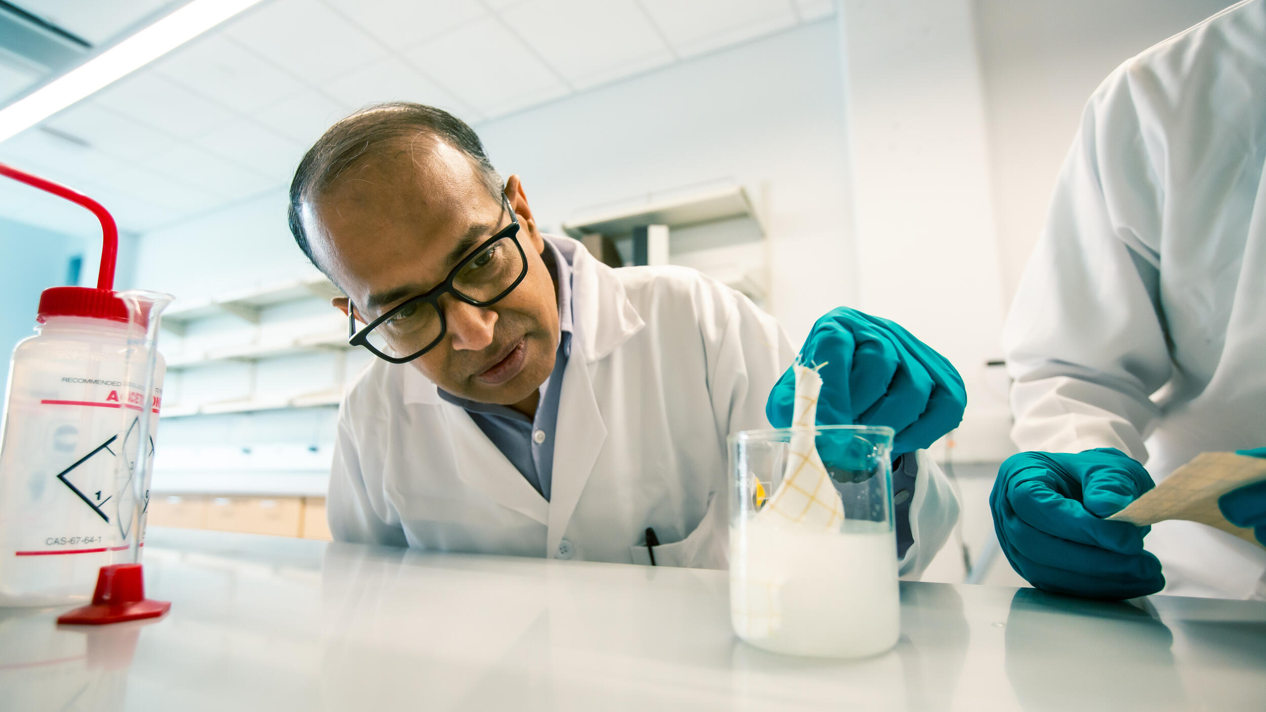 A photo of a man putting a baloon in a beaker filled with a white liquid. 