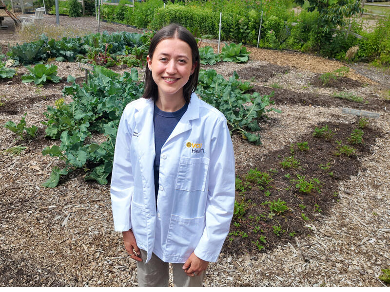 A photo of a woman wearing a white medical coat that says \"VCU Health\" standing in a vegtable garden. 