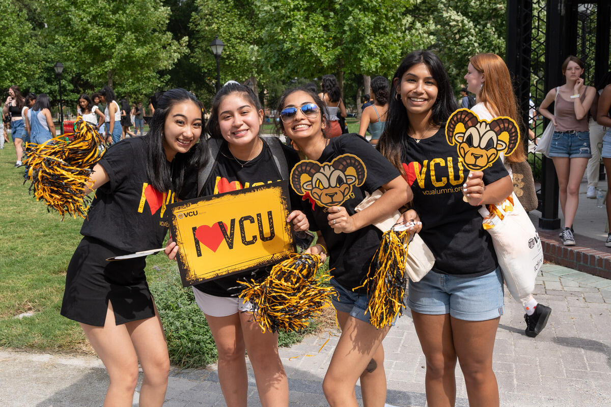 A photo of four women stading next to eachother holding pom poms, a sign that says \"I heart VCU,\" and paper rodney the ram masks. 
