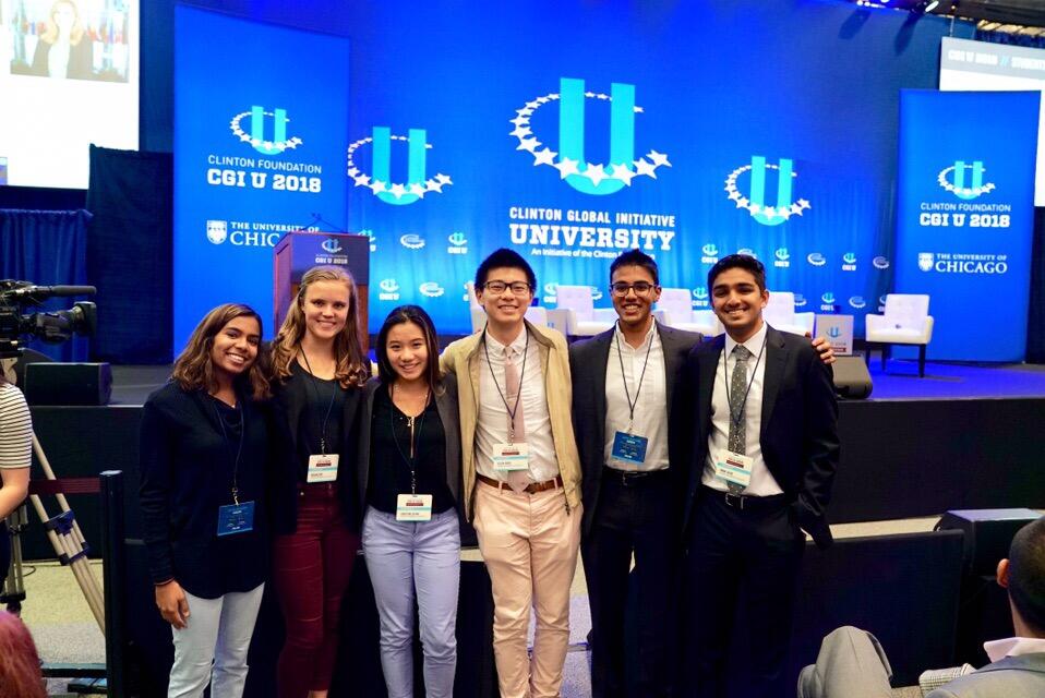 A group of six people in business attire stand in front of signs reading "Clinton Global Initiative University."