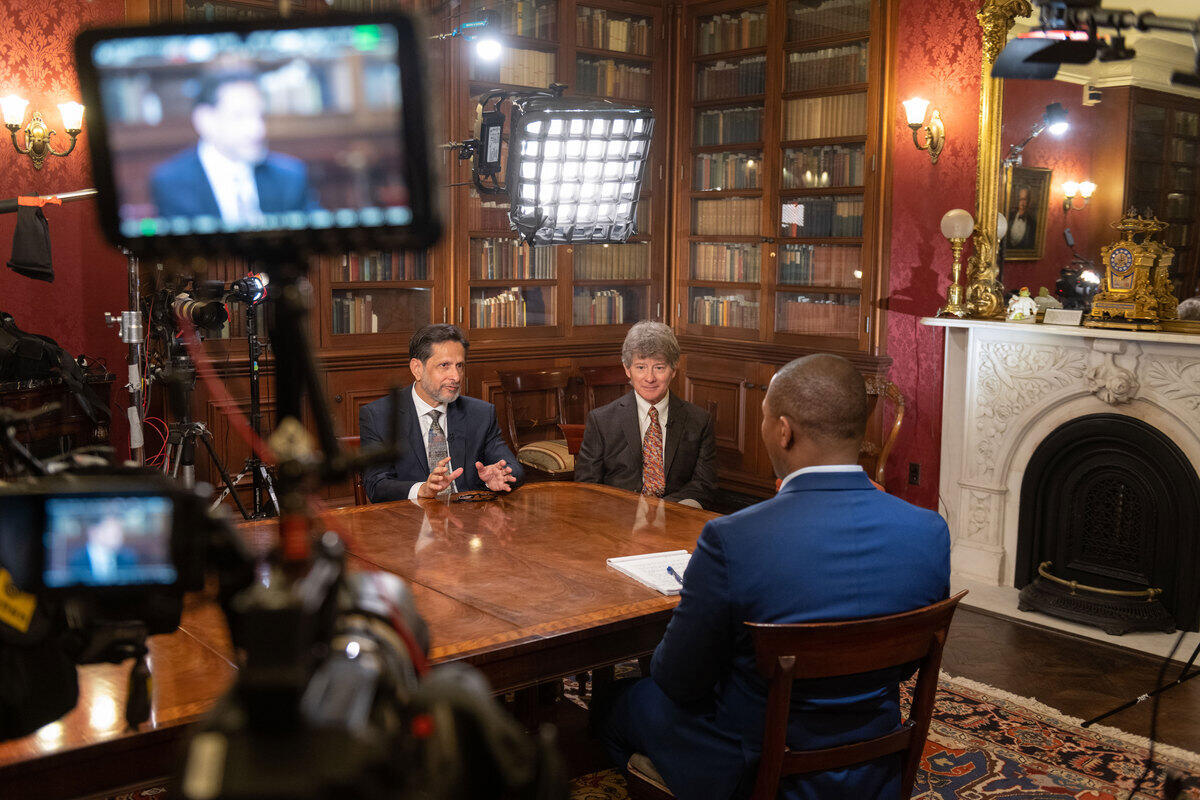 Two men sit on one side of a table in a library while a journalist sits across from them with his back to the camera. A light sits behind the men being interviewed, while a video camera in the foreground captures the image of one interviewee who is speaking.