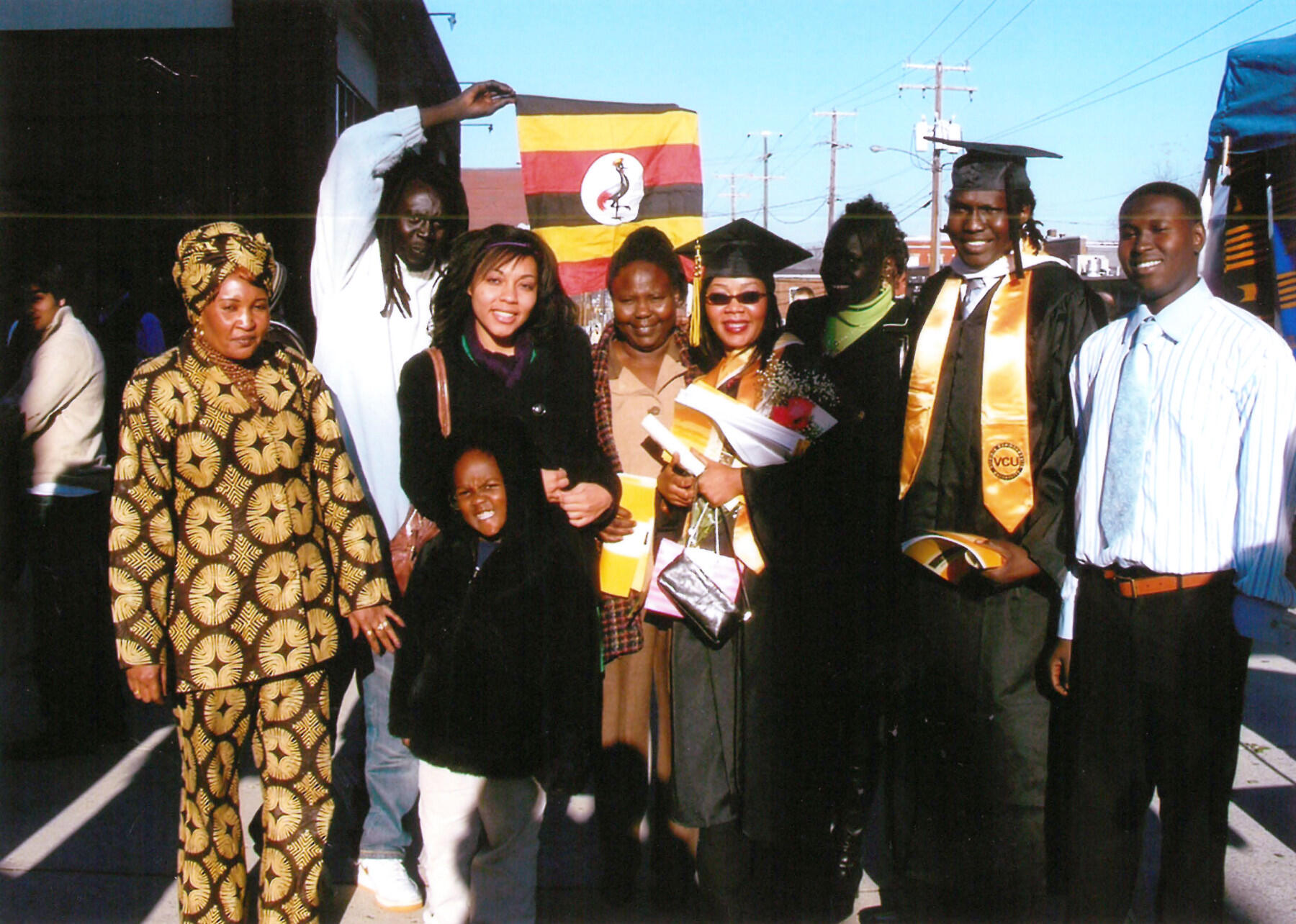 A group of people in dress clothes and graduation robes stands outside. One man holds a red, yellow and black (Ugandan) flag.