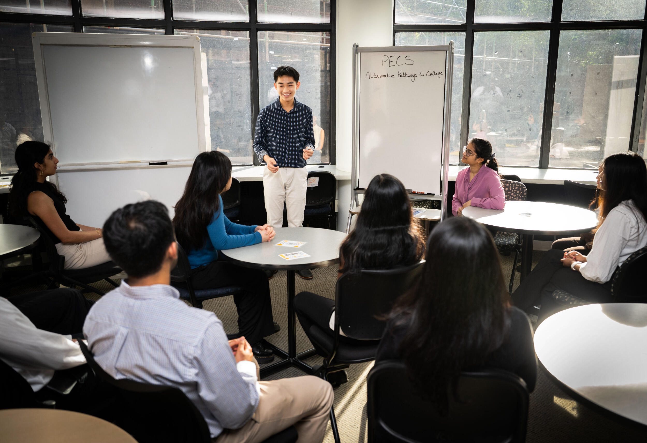 A man stands at the front of a room in front of a white board and men and women sit in chairs around him watching him speak.