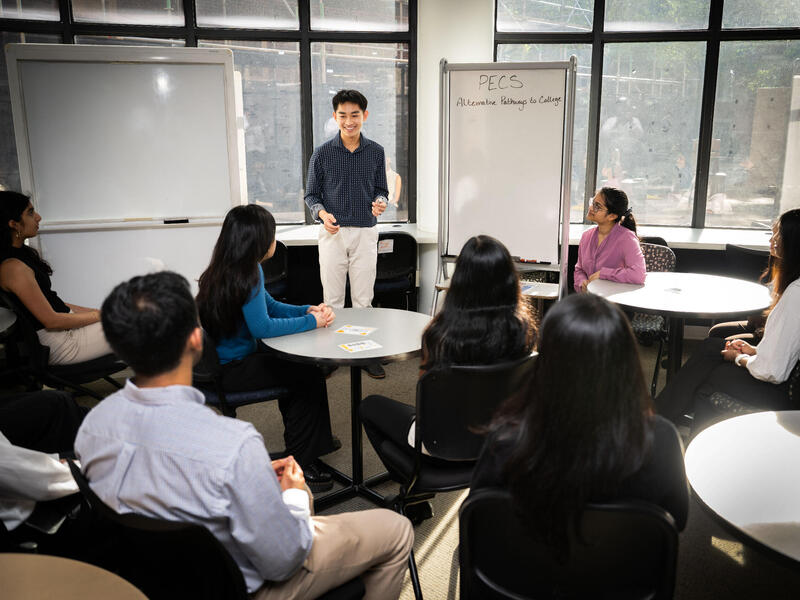 A man stands at the front of a room in front of a white board and men and women sit in chairs around him watching him speak.