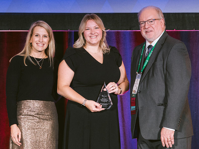 A photo of three people standing. The woman in the middle is holding a glass trophy. 