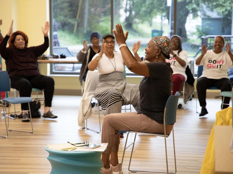 A photo of a room full of women sitting in chairs and raising their arms above their heads. 