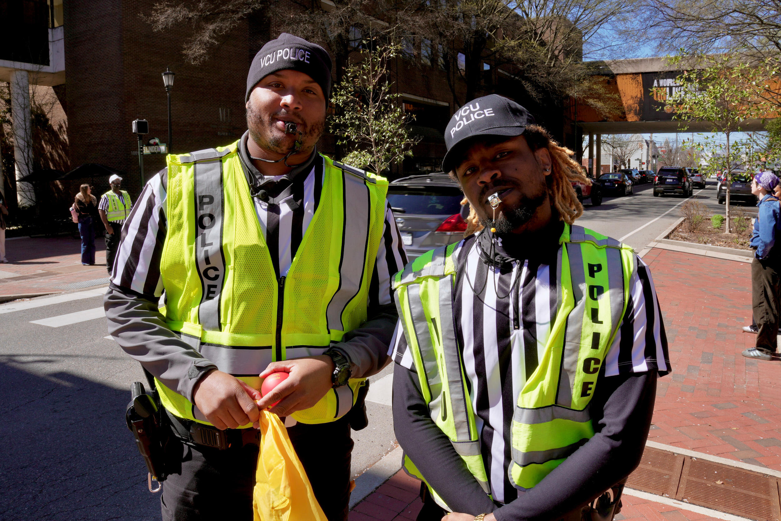 Two police officers in striped referee shirts and reflective vests pose with whistles in their mouths.
