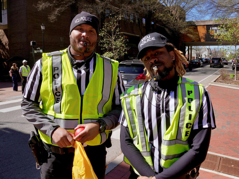 Two police officers in striped referee shirts and reflective vests pose with whistles in their mouths.