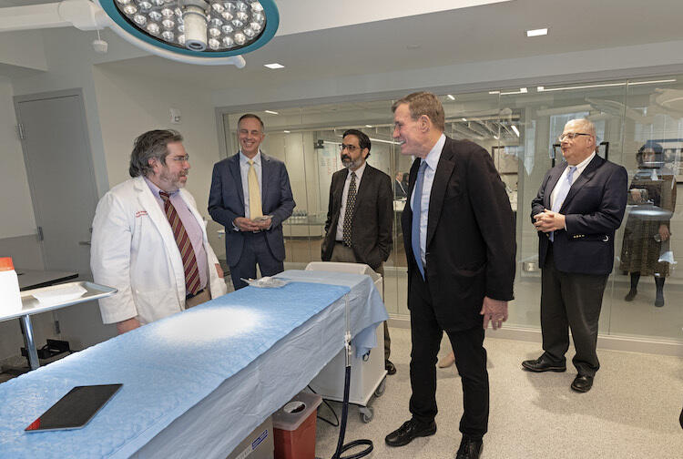 A photo of five men standing in a medical proceedure room, in a semi-circle around a medical bed. The man on the far left is wearing a white doctor's coat. The other four men are wearing suits and ties. 