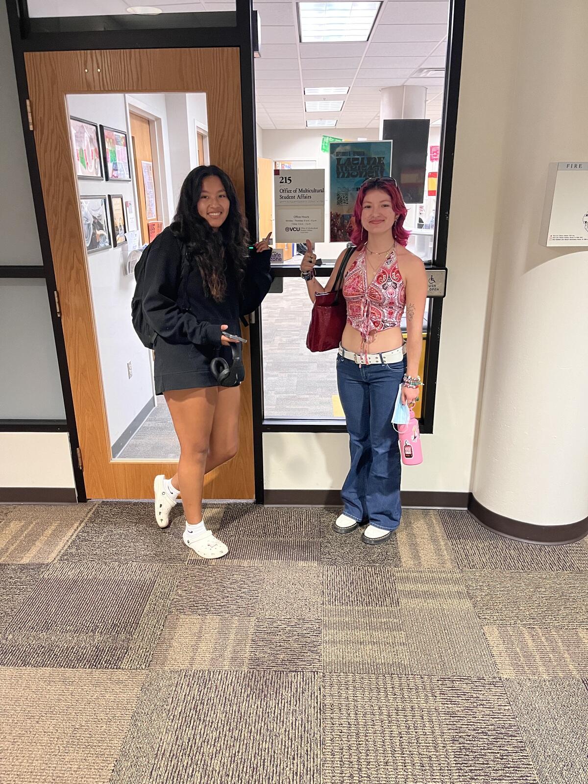 Two women standing in front of a door poitning at a sign that says \"Office of Multicultural Affairs\" 